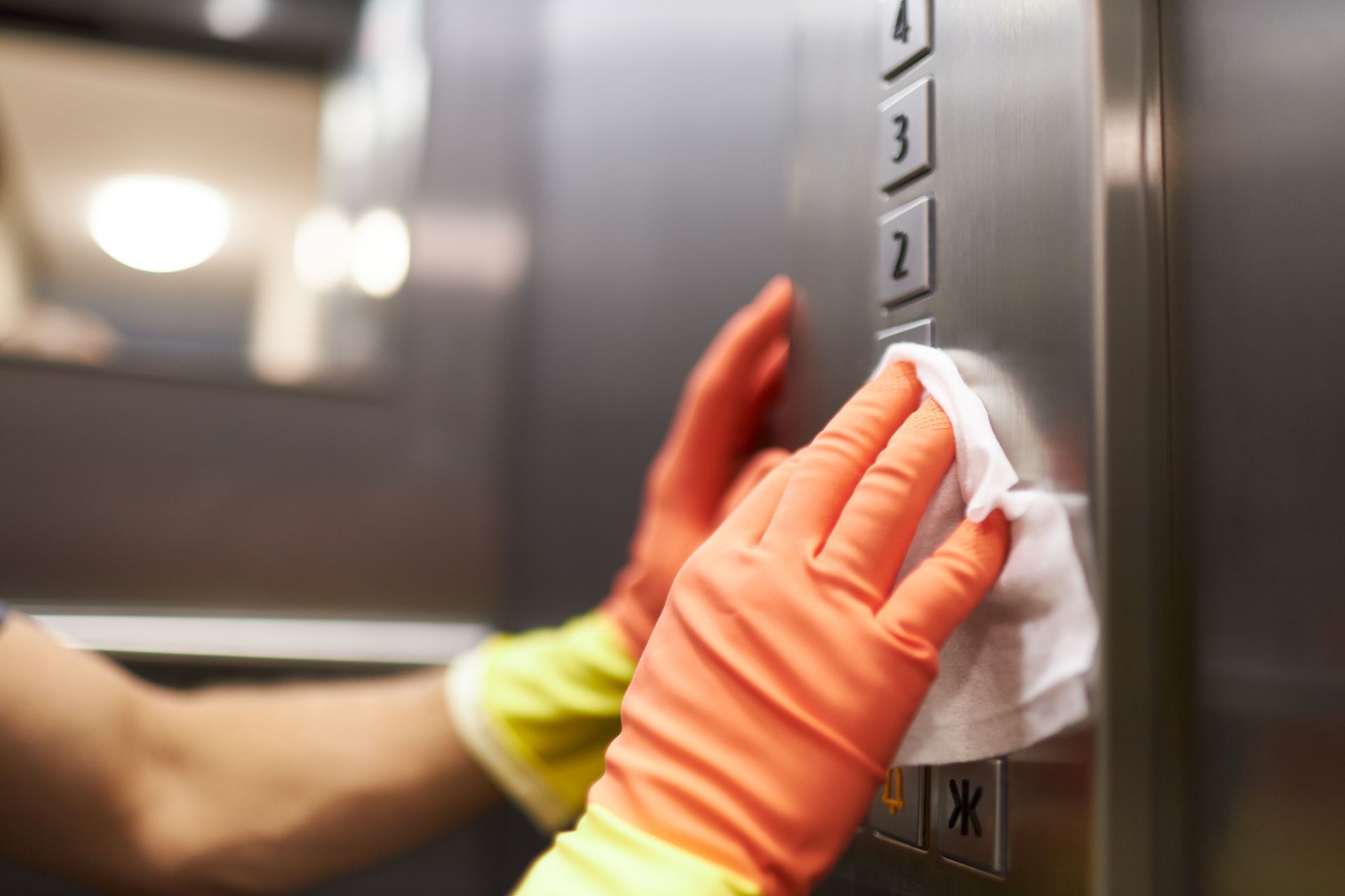 Person wearing orange gloves wiping elevator buttons with a white cloth.