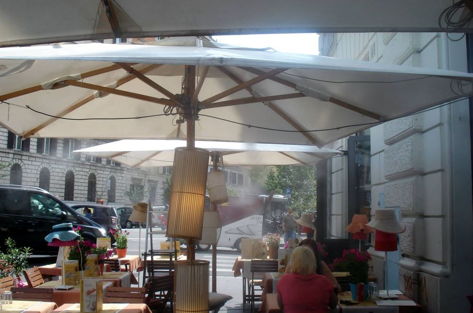 Tavolini all'aperto di un caffè sotto un grande ombrellone bianco, con auto parcheggiate e una persona con una maglietta rosa seduta sulla destra.