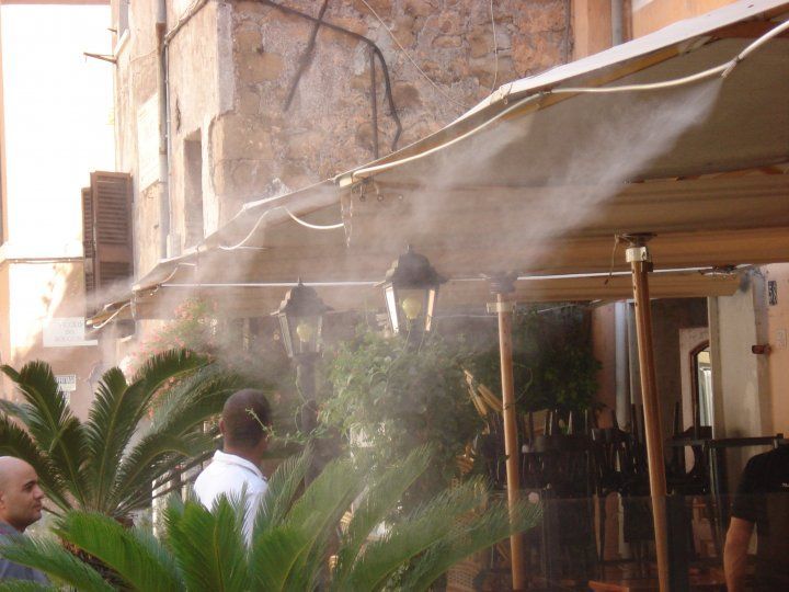 Persone che camminano accanto a ventilatori nebulizzatori esterni, vicino a un edificio in pietra e a una tettoia.