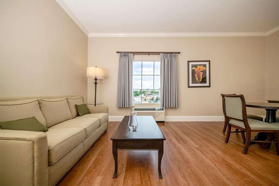 Living room with couch, table, chairs, window, and artwork. Beige walls and hardwood floors.