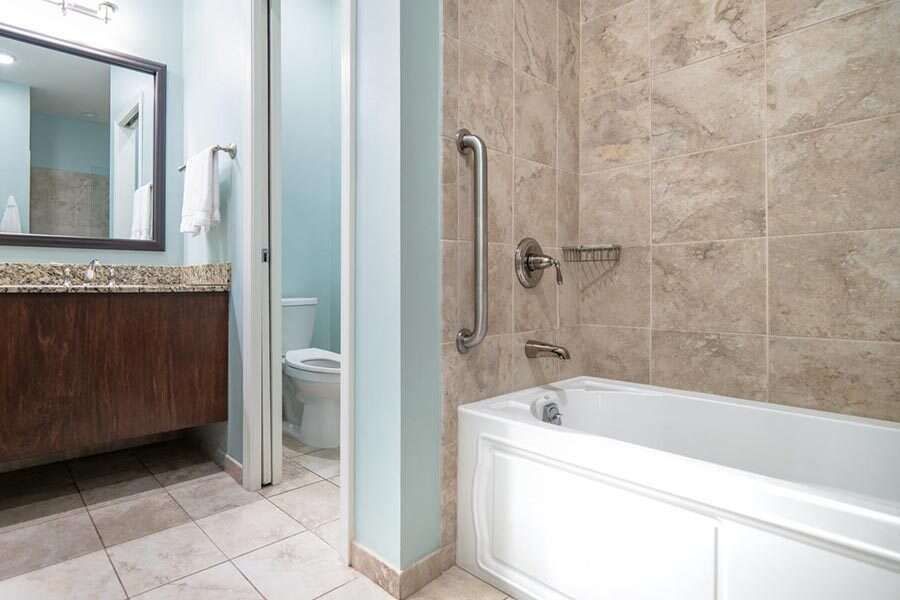 Bathroom with bathtub, toilet, and sink. Light blue walls, beige tiles, and wood vanity.