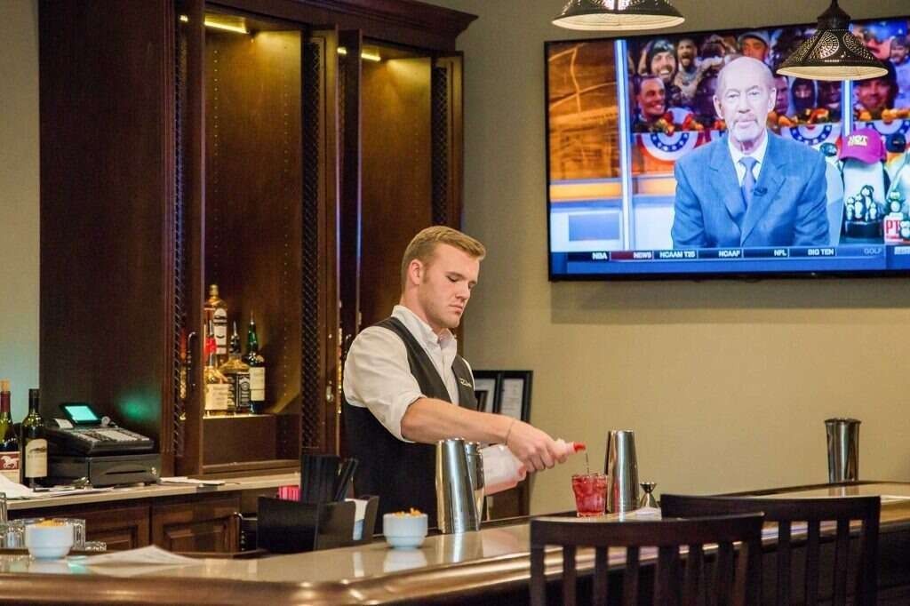 Bartender pouring ingredients into a shaker at a bar, TV in background showing a news report.