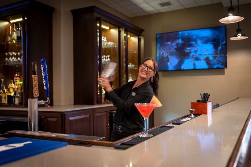 Bartender smiles while shaking a cocktail behind a bar, television overhead.