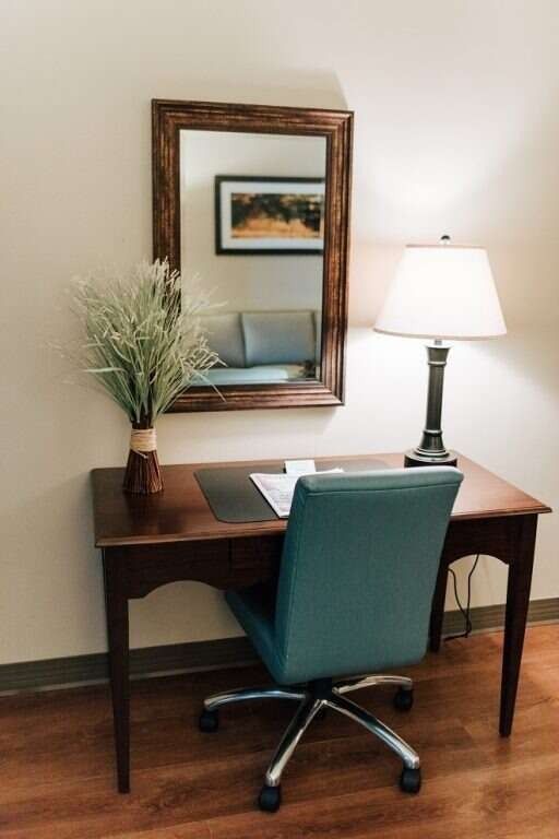 Desk with chair, lamp, and mirror. Green chair faces desk. Mirror reflects couch and artwork. Light brown wall.