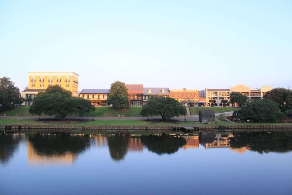 Skyline of buildings reflected in calm water, green trees along the shore.