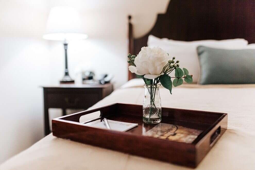 A flower and card on a tray on a made bed in a hotel room with a nightstand and lamp.