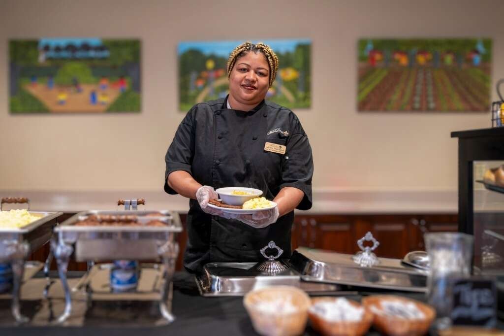 Chef in black uniform holding a plate of food, smiling, behind a buffet table with food containers, artwork on the wall.