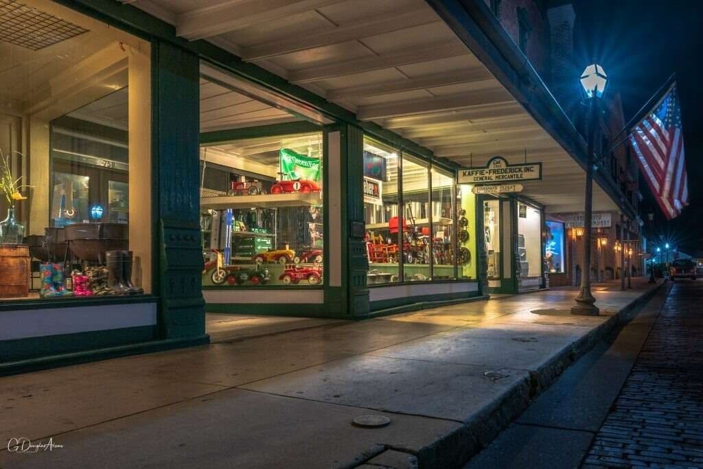 Night view of storefronts on a brick-paved street, with American flags and illuminated displays.