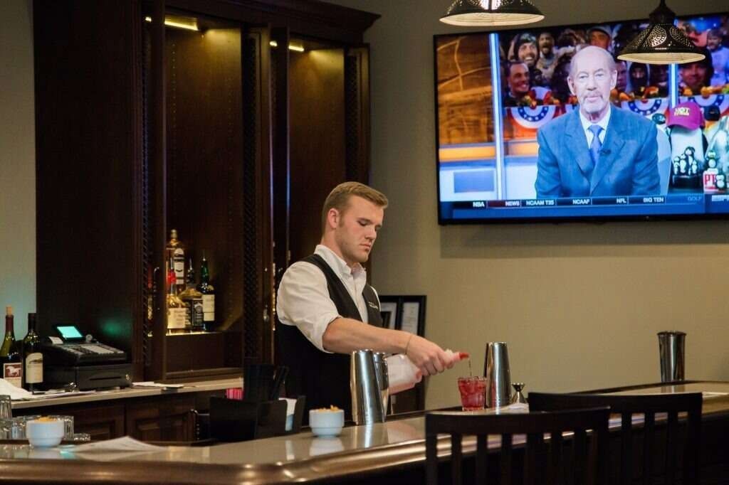 Bartender mixing drinks behind a bar, TV screen in background showing a sports broadcaster.