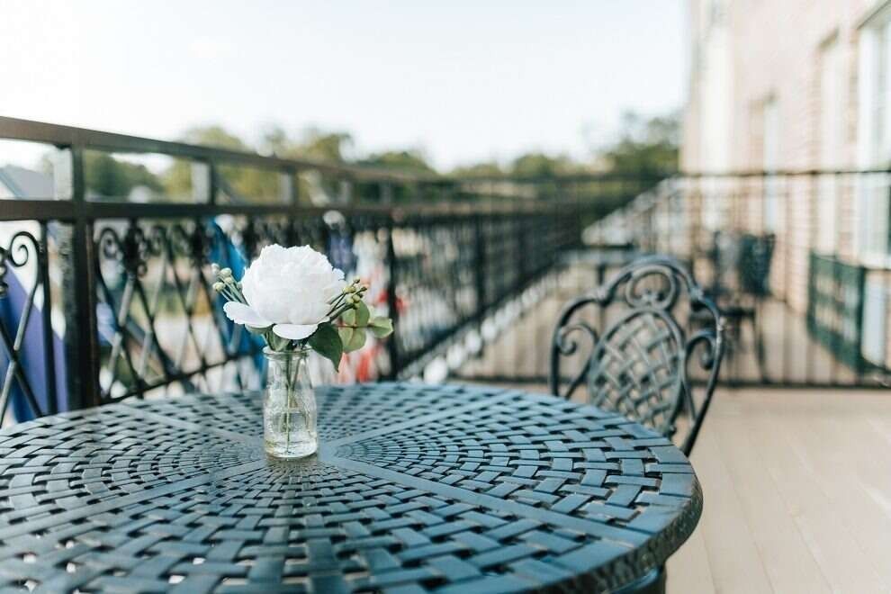 Black patio table with a vase of white flowers on a balcony overlooking a sunny vista.
