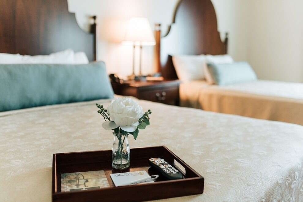 Hotel room with two beds; white floral arrangements on a tray, aqua pillows, nightstand with lamp, and light bedding.