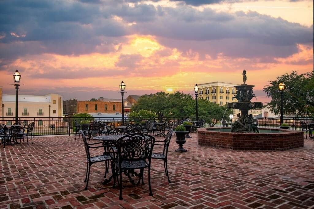 A brick-paved plaza with iron tables, a fountain, and buildings at sunset.