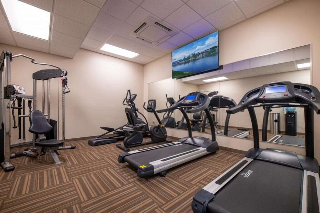 Gym with exercise machines, TV, and large mirror. Brown patterned carpet and neutral walls.