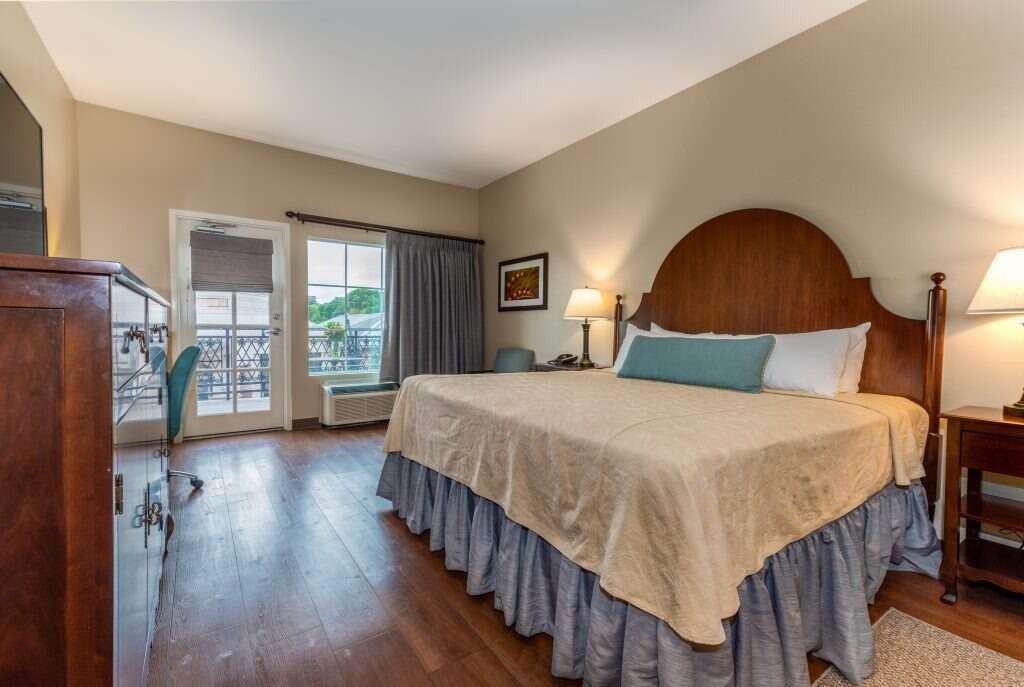 Hotel bedroom with king-size bed, wooden furniture, balcony access, and neutral colors.