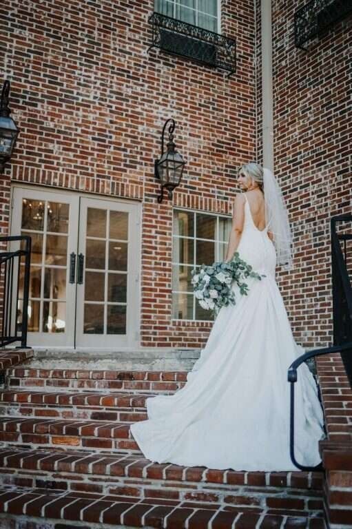 Bride in white dress and veil poses on brick steps, holding a bouquet, near building with glass doors.