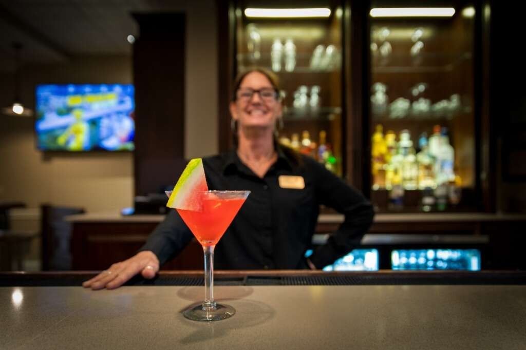 Bartender smiles behind the bar with a pink cocktail garnished with a watermelon slice.
