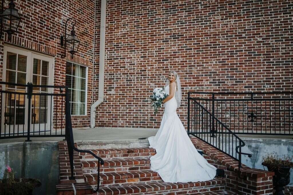 Bride in white dress holding flowers, on brick steps, in front of a brick building.