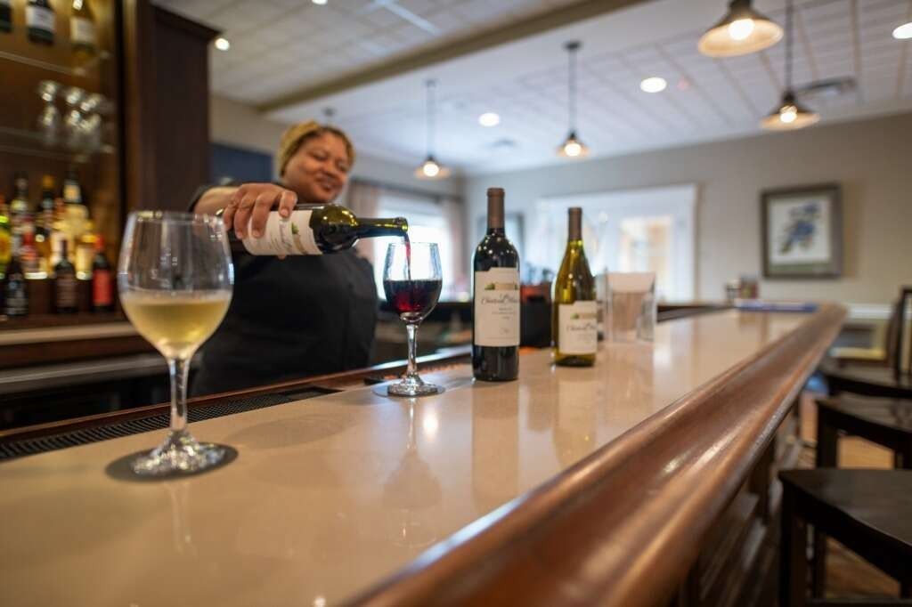 Bartender pours red wine into a glass at a bar. Two wine bottles sit nearby.
