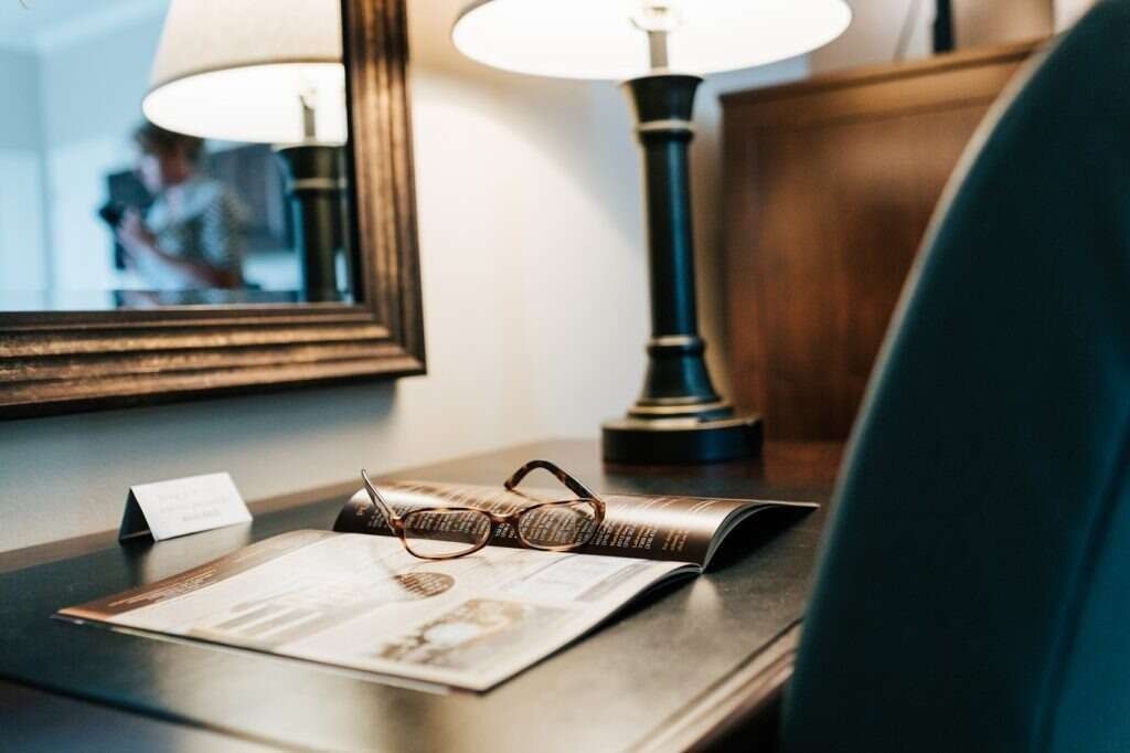 Desk with lamp, mirror, open magazine, glasses. Person in reflection.