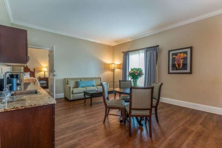 Spacious hotel suite with living area, dining table, and view of bedroom. Dark wood floors, beige walls.