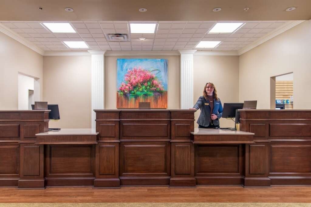 Woman behind a wooden bank teller counter with a colorful painting, in a well-lit room.