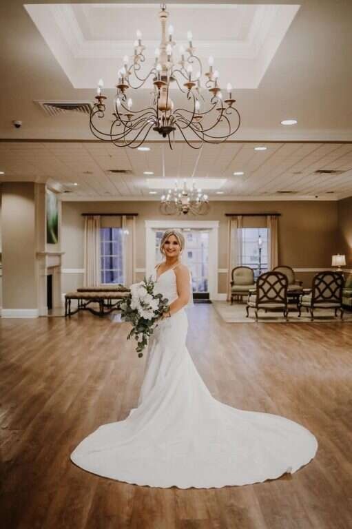 Bride in a white gown holding bouquet, standing in a wedding reception hall with chandelier.