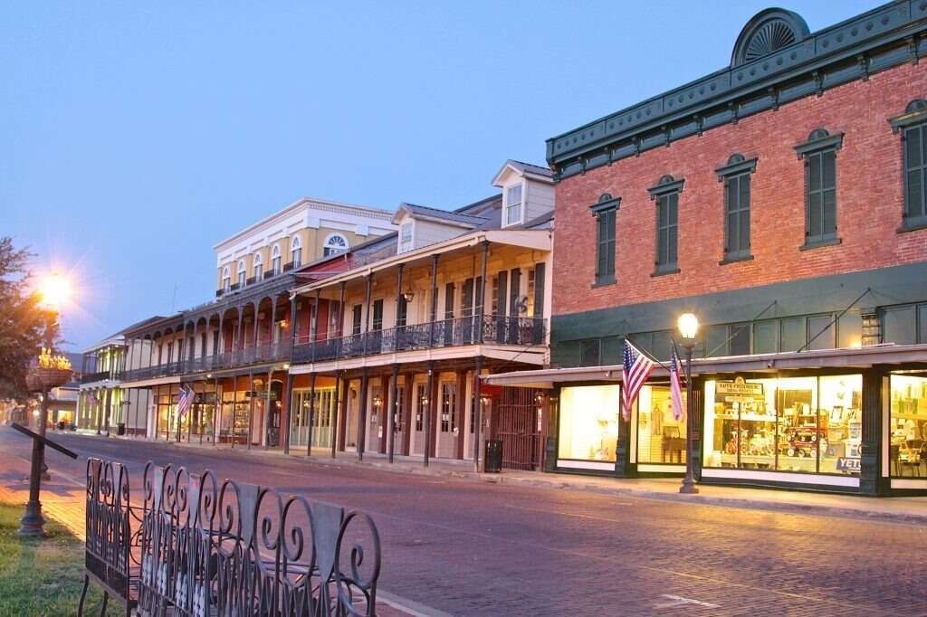 Street with historic brick buildings, shop fronts, and a wrought-iron fence in a town at dusk.