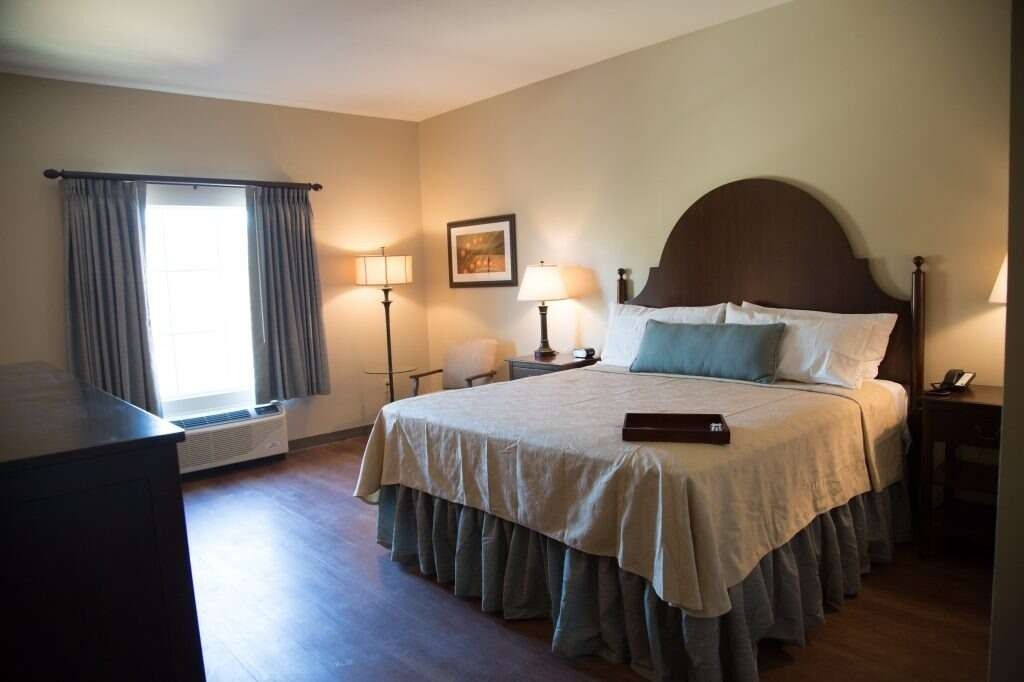 Hotel room with king bed, lamps, window with curtains, dresser, and art.