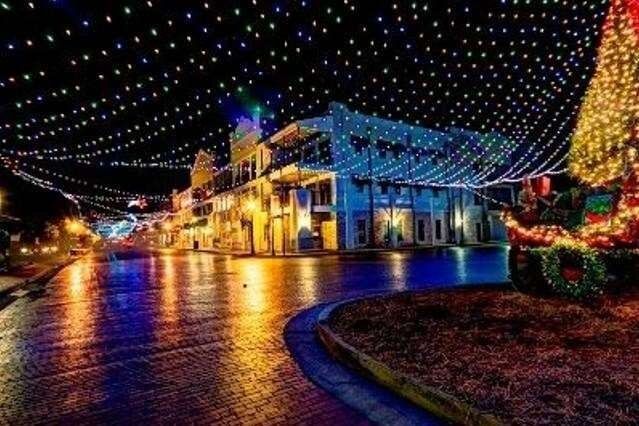 Nighttime street decorated with Christmas lights reflecting on a wet street. Buildings are lit up with a festive tree.