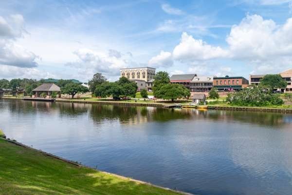 Calm river scene with buildings and trees under a partly cloudy sky.