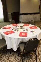 Round table with red folders, surrounded by black chairs, in a room with a projector screen.