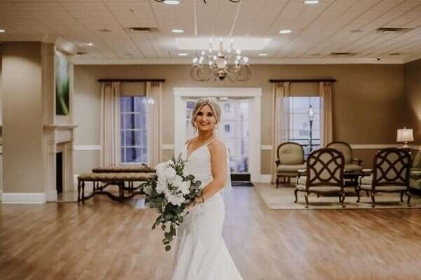 Bride in white dress holding bouquet, standing in a well-lit reception hall with neutral tones.