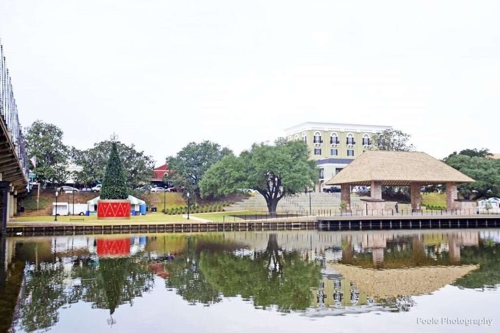 Waterfront park with reflections of trees, buildings, and a gazebo in the water under an overcast sky.