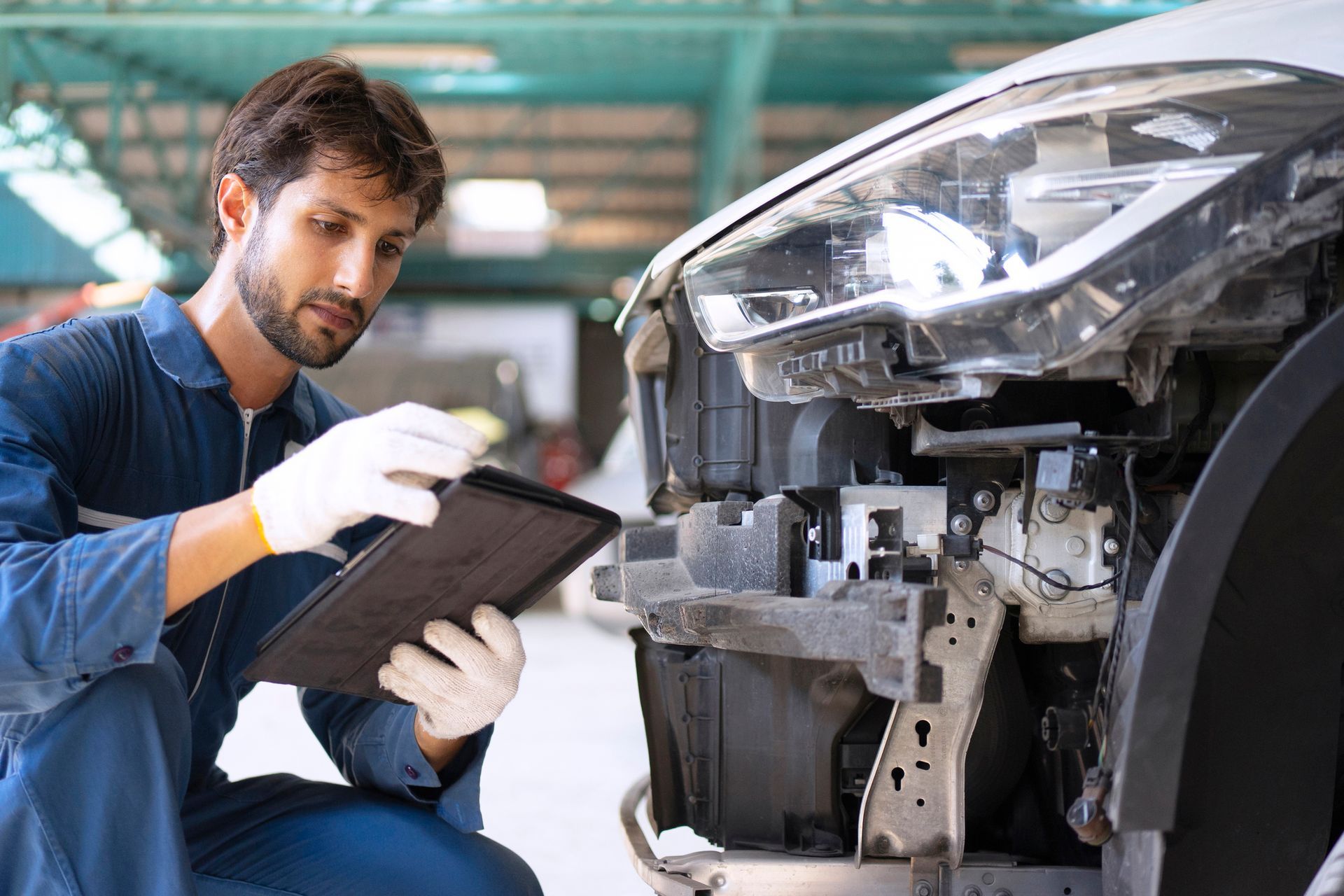 Mechanic in blue overalls and gloves examines a car's damaged front end, holding a tablet.