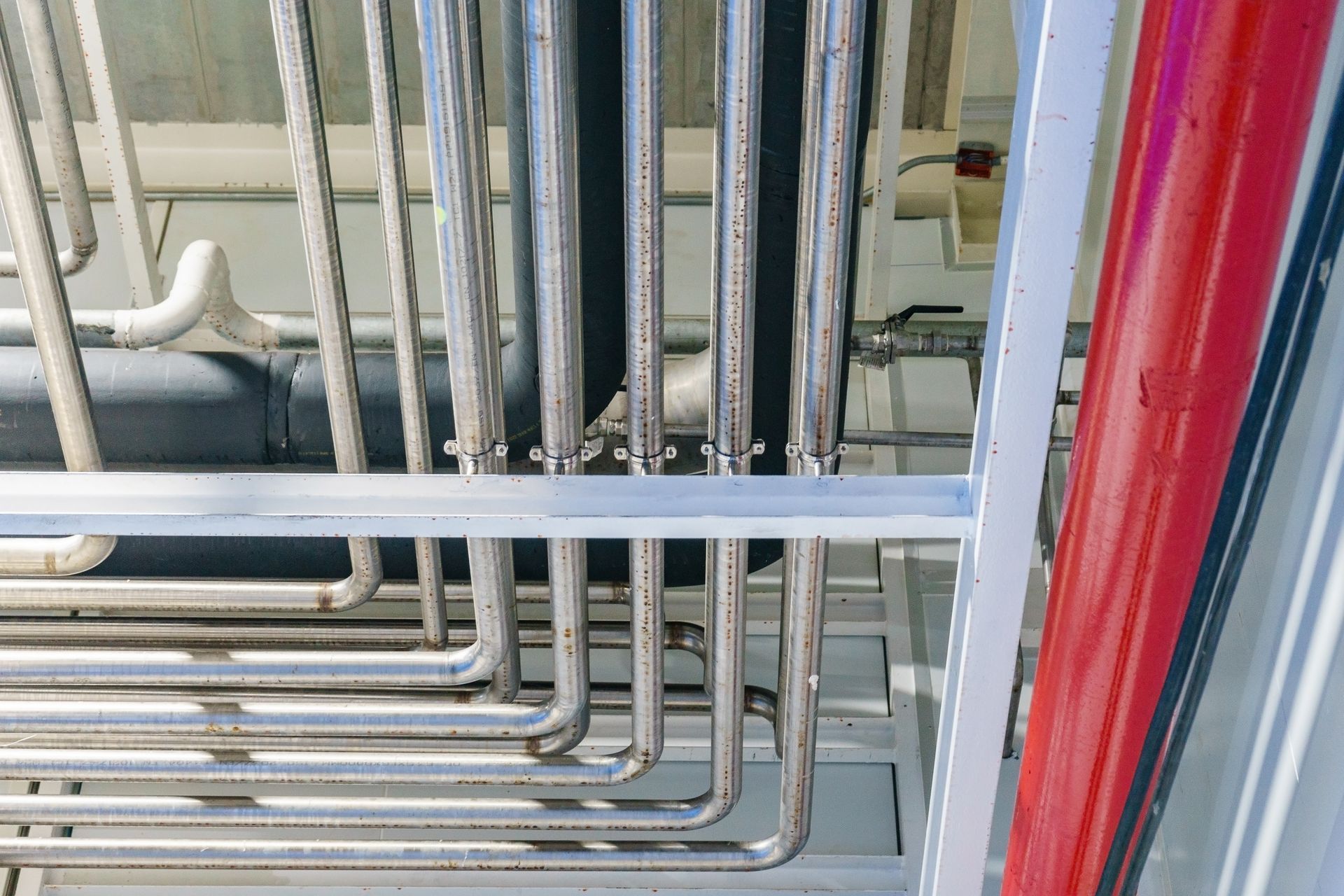 Industrial pipes and conduit running vertically and horizontally in a utility room, with red and gray piping.