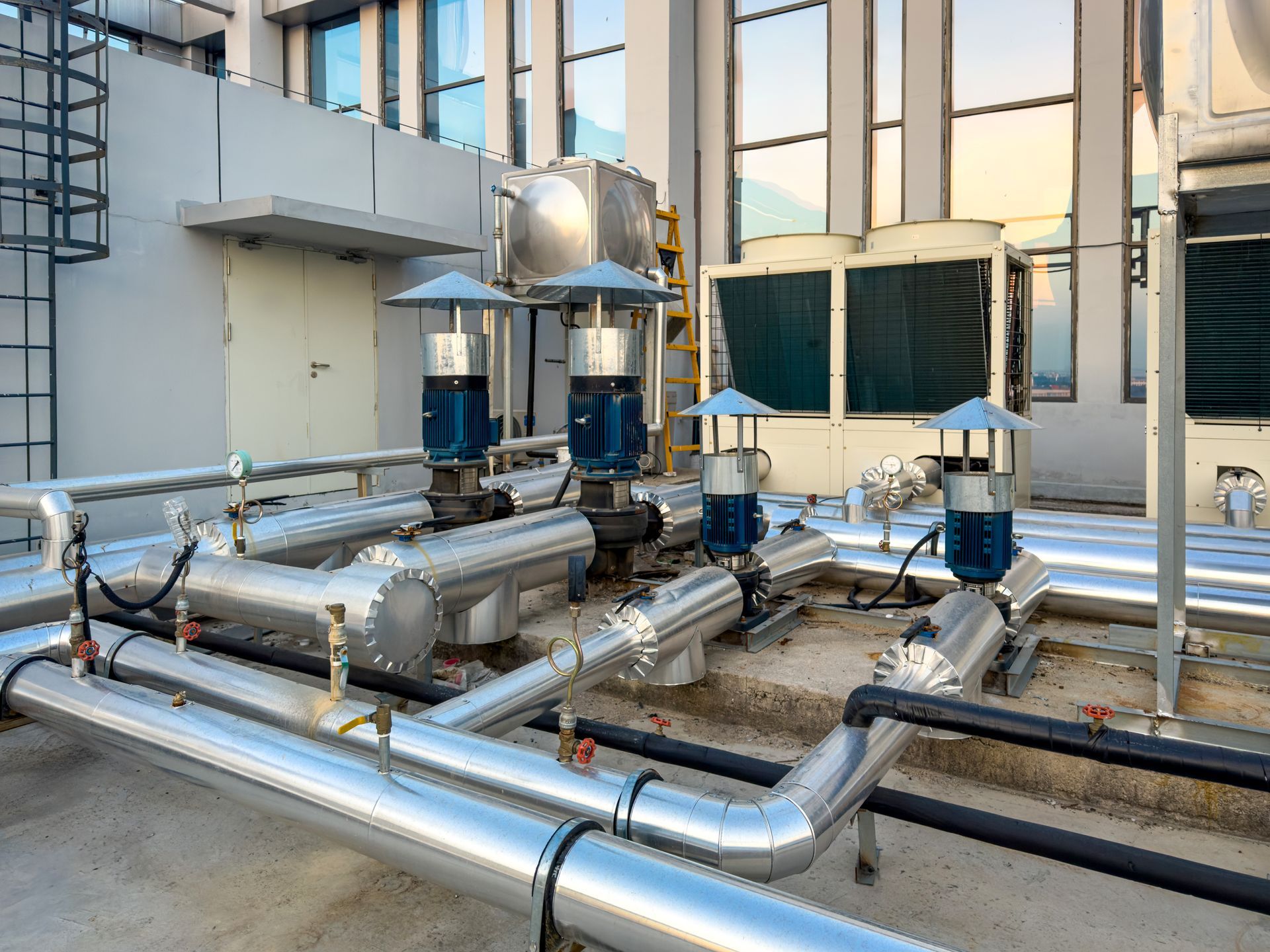 Rooftop HVAC piping and equipment beside a modern building, with shiny metal ducts and blue ventilation units.