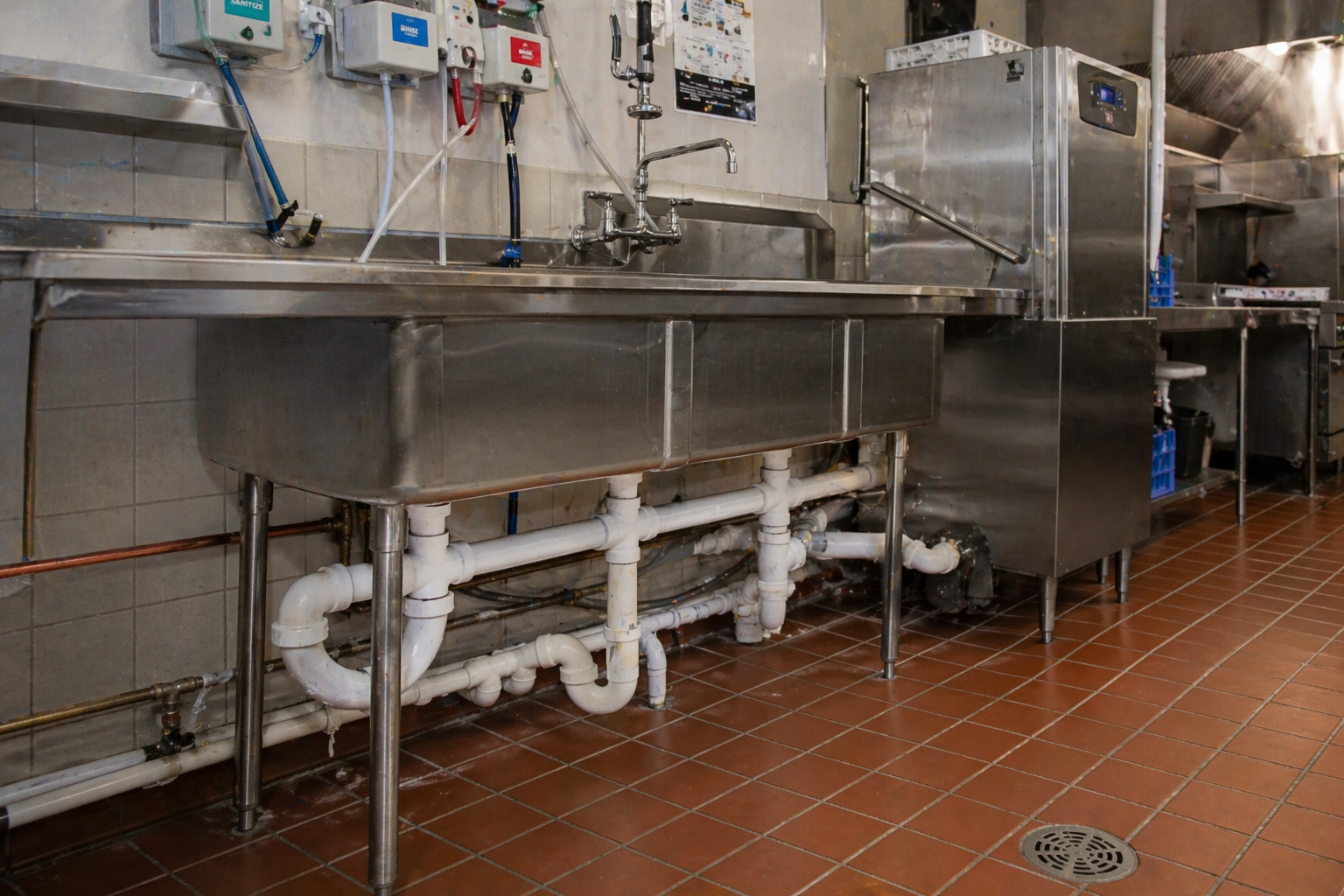 Technician kneels under a stainless-steel sink, repairing pipes in a commercial kitchen.