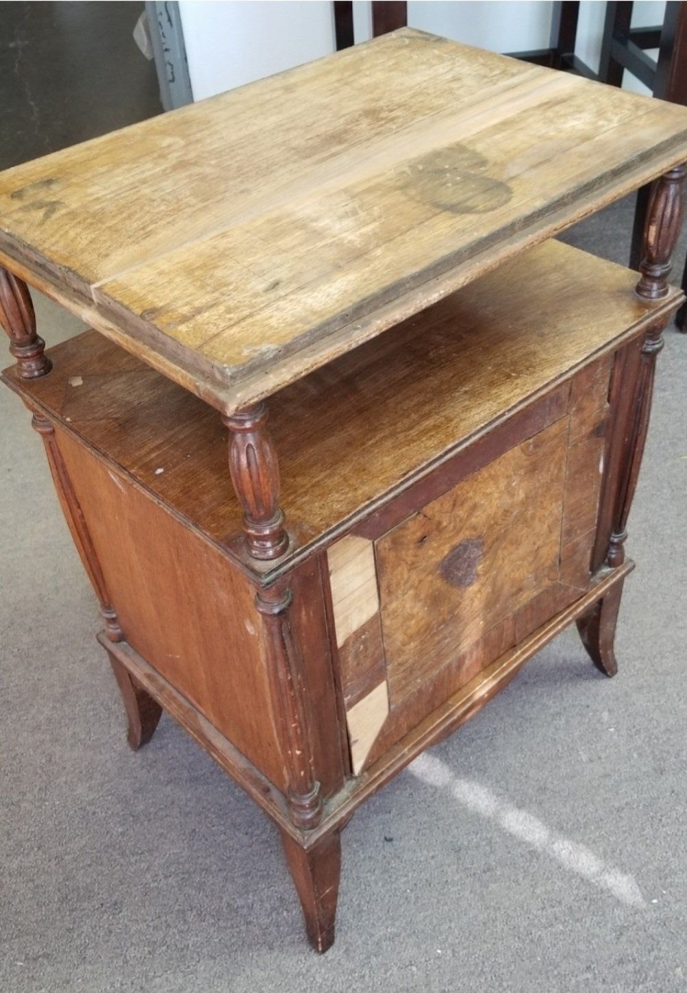 A weathered, wooden two-tiered side table with spindled legs and a missing piece of veneer on the front cabinet door.