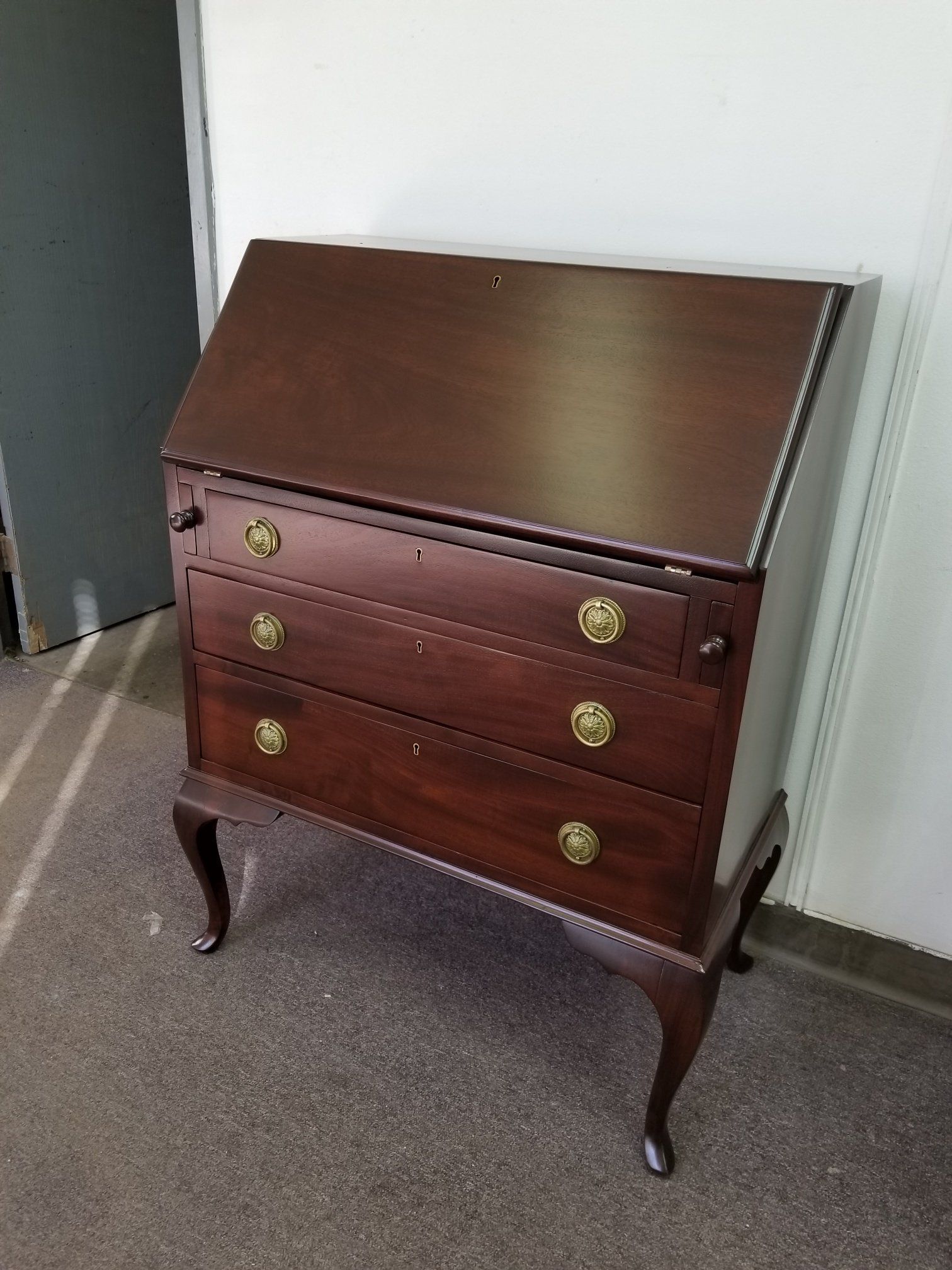 Mahogany wooden secretary desk with brass handles and curved legs against a white wall.