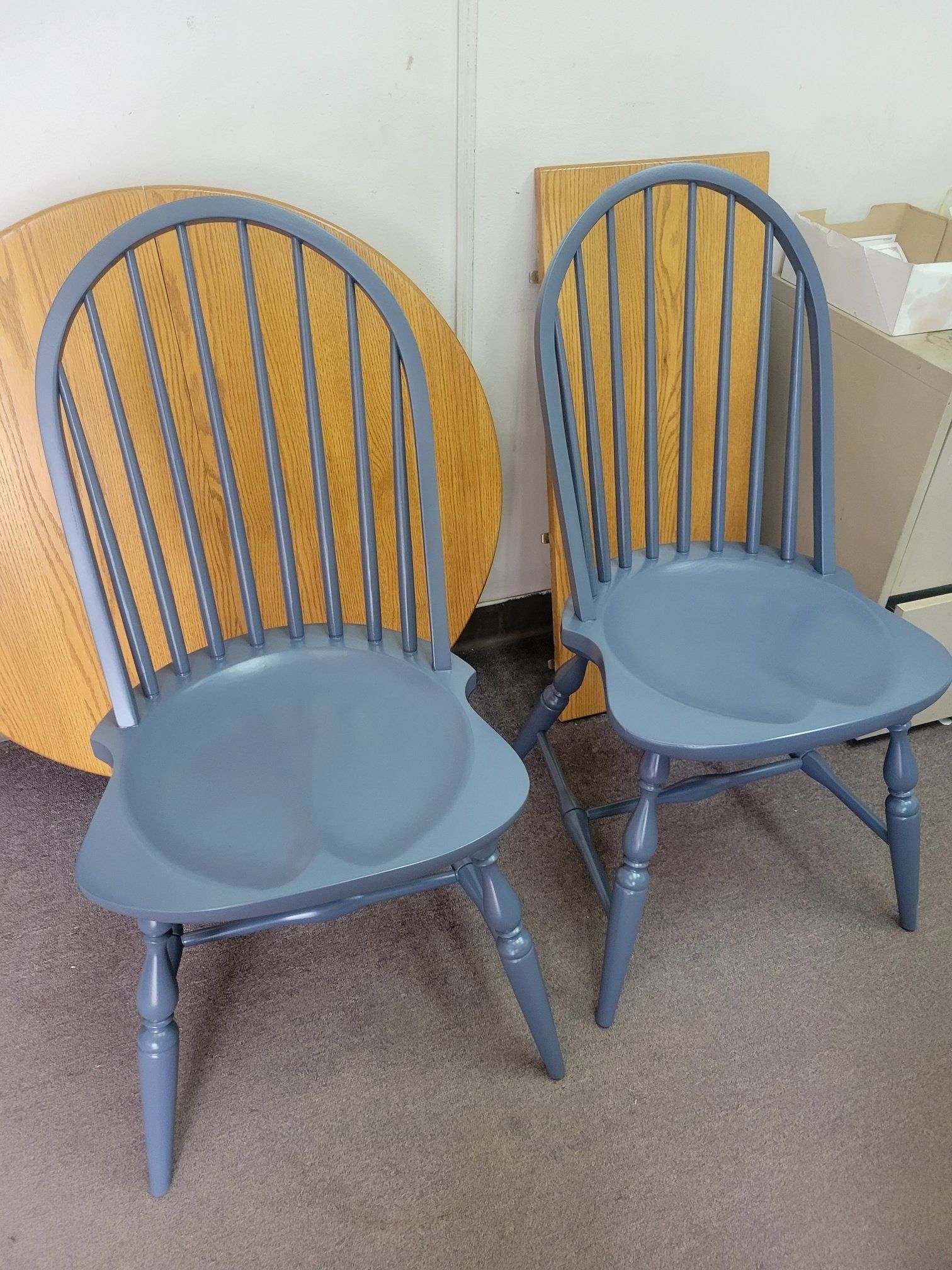 Two blue wooden chairs in a room, with round tabletops leaning behind them.
