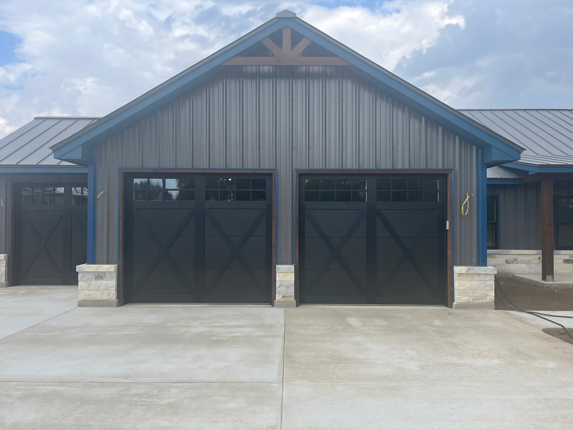 Gray metal barn with three black garage doors and a concrete driveway under a cloudy sky