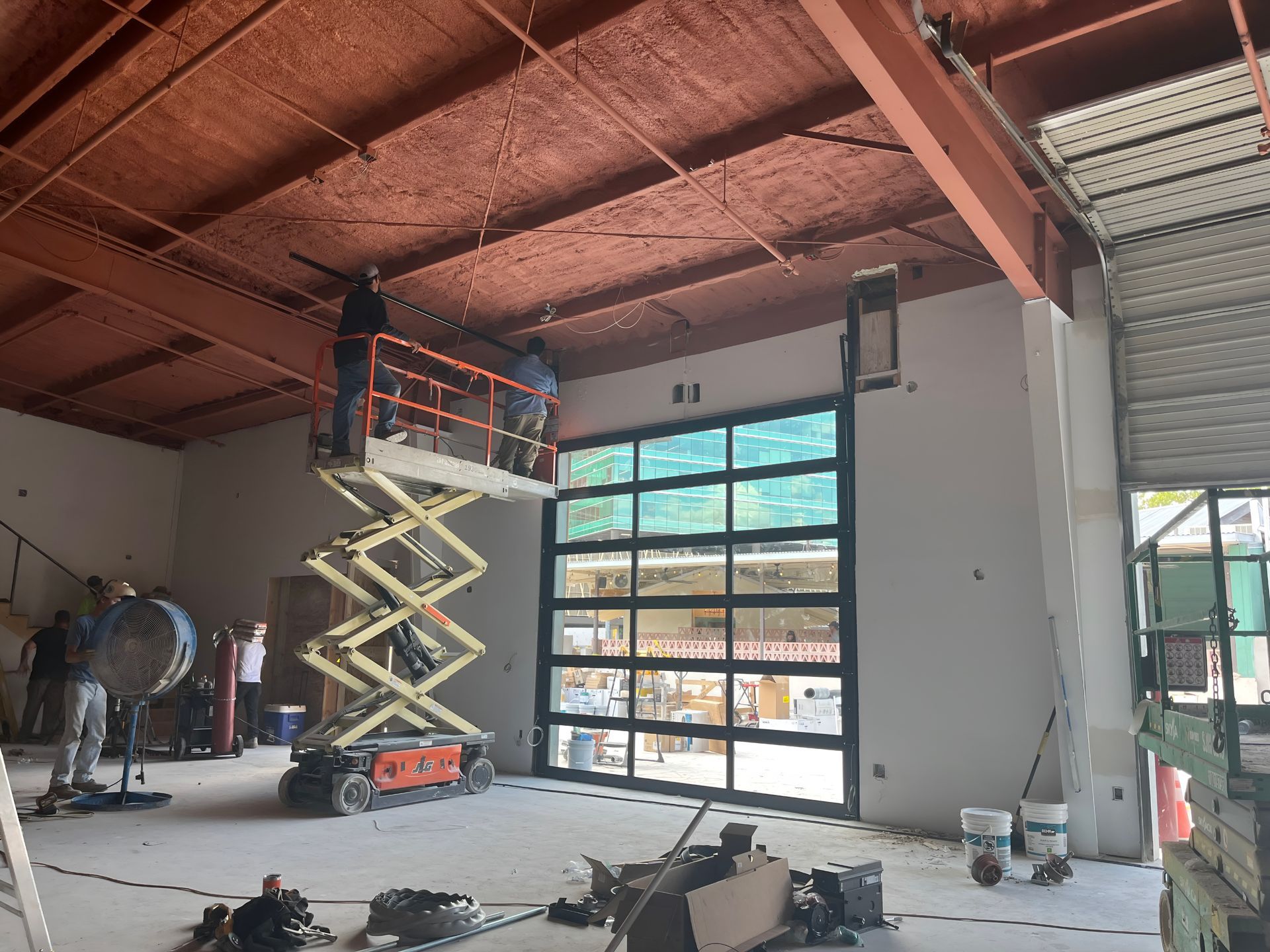 Worker on a scissor lift installing a large industrial window inside a warehouse under construction.