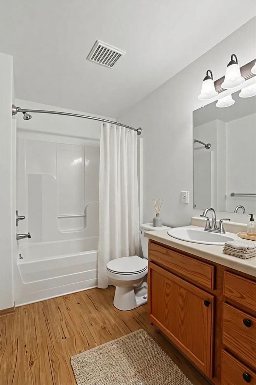 Bright bathroom with white tub and shower curtain, toilet, wood vanity, mirror, and beige rug.