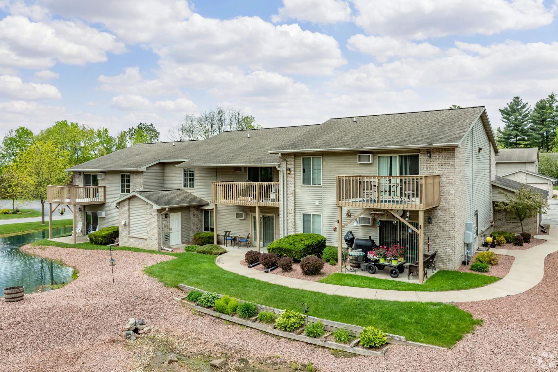 Apartment complex with tan siding, balconies, and a landscaped gravel courtyard beside a pond