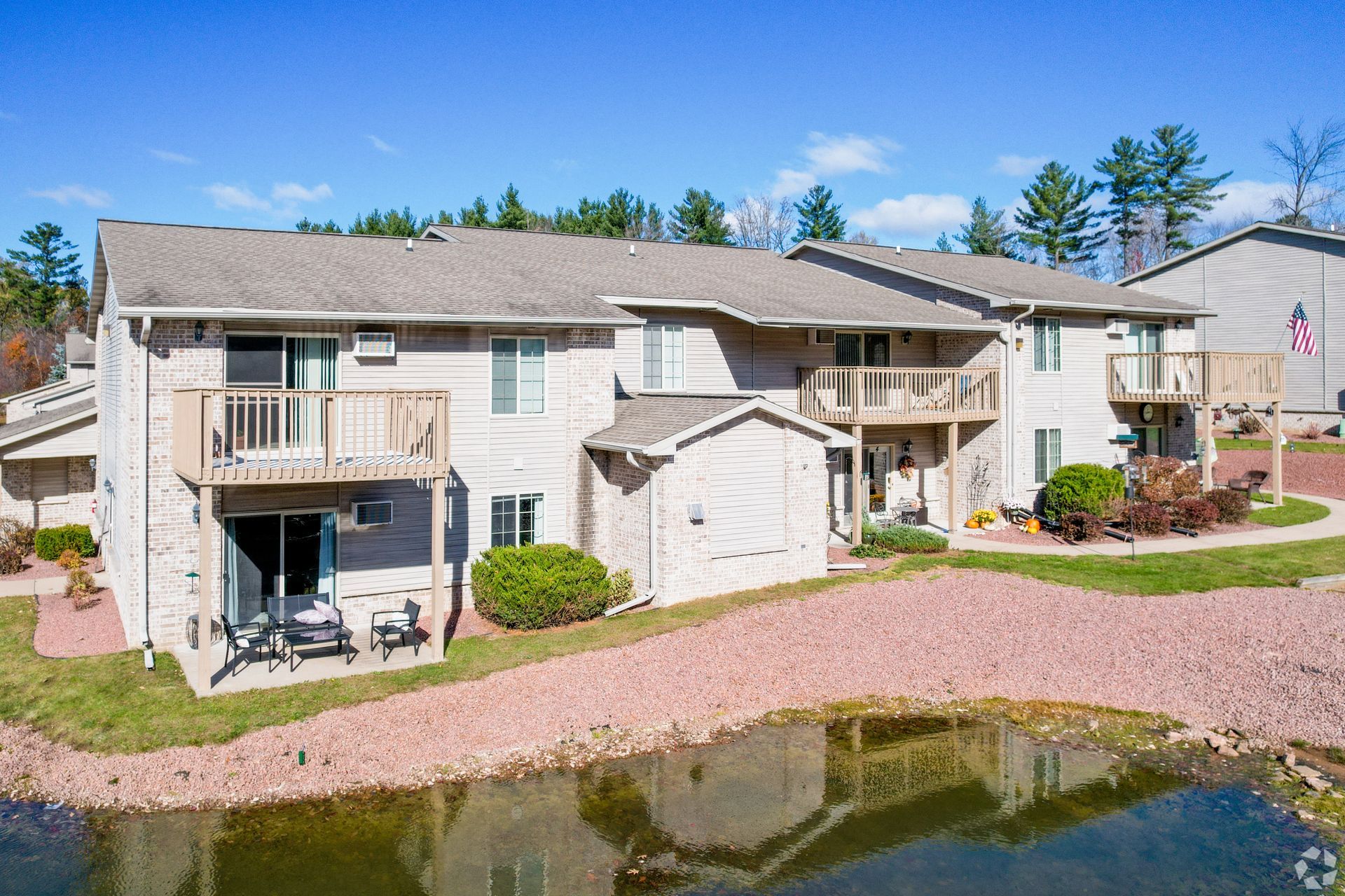 Apartment buildings beside a pond, with balconies and a gravel path under a clear blue sky