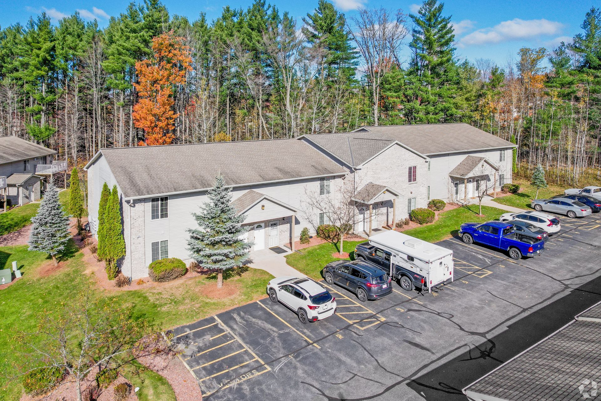 Aerial view of a white apartment complex with parked cars and a wooded backdrop.