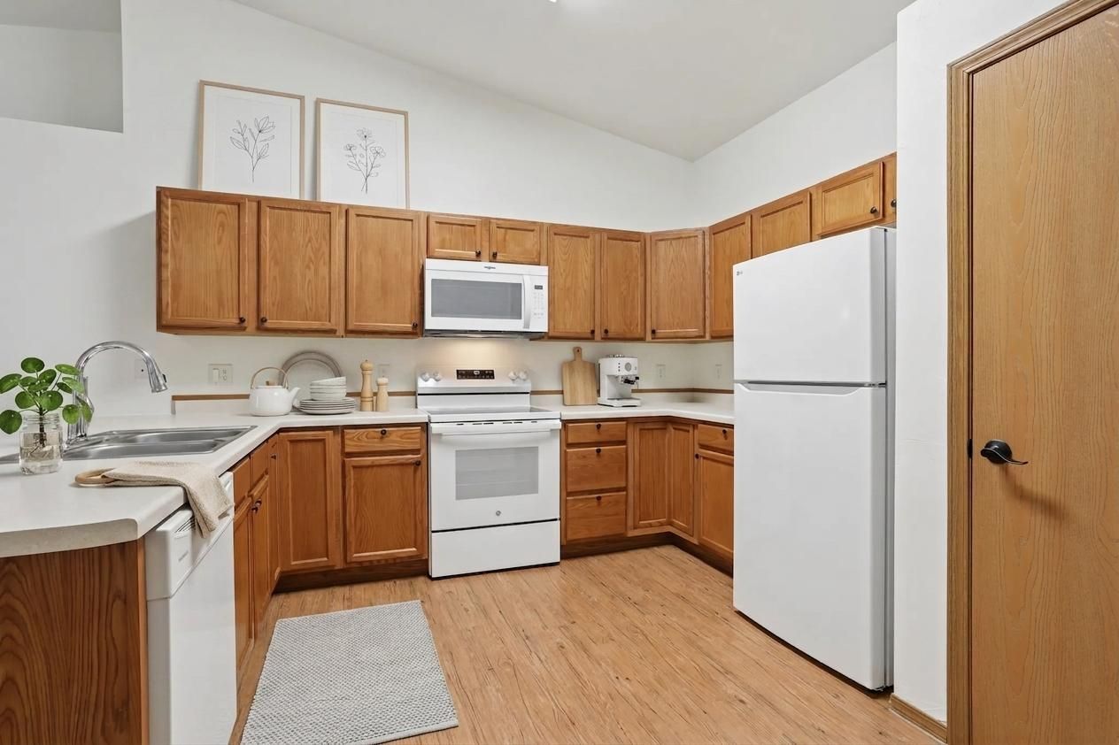 Compact kitchen with wooden cabinets, white appliances, sink, stove, microwave, and a beige rug on the floor.