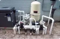 Pool equipment pipes and valves beside a beige filter tank outdoors, near a gray utility box.