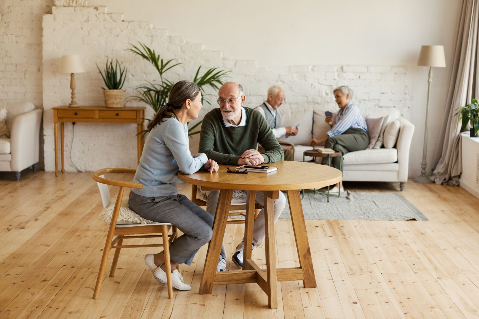 A young woman and elderly man play dominoes at a table in a room with other people.