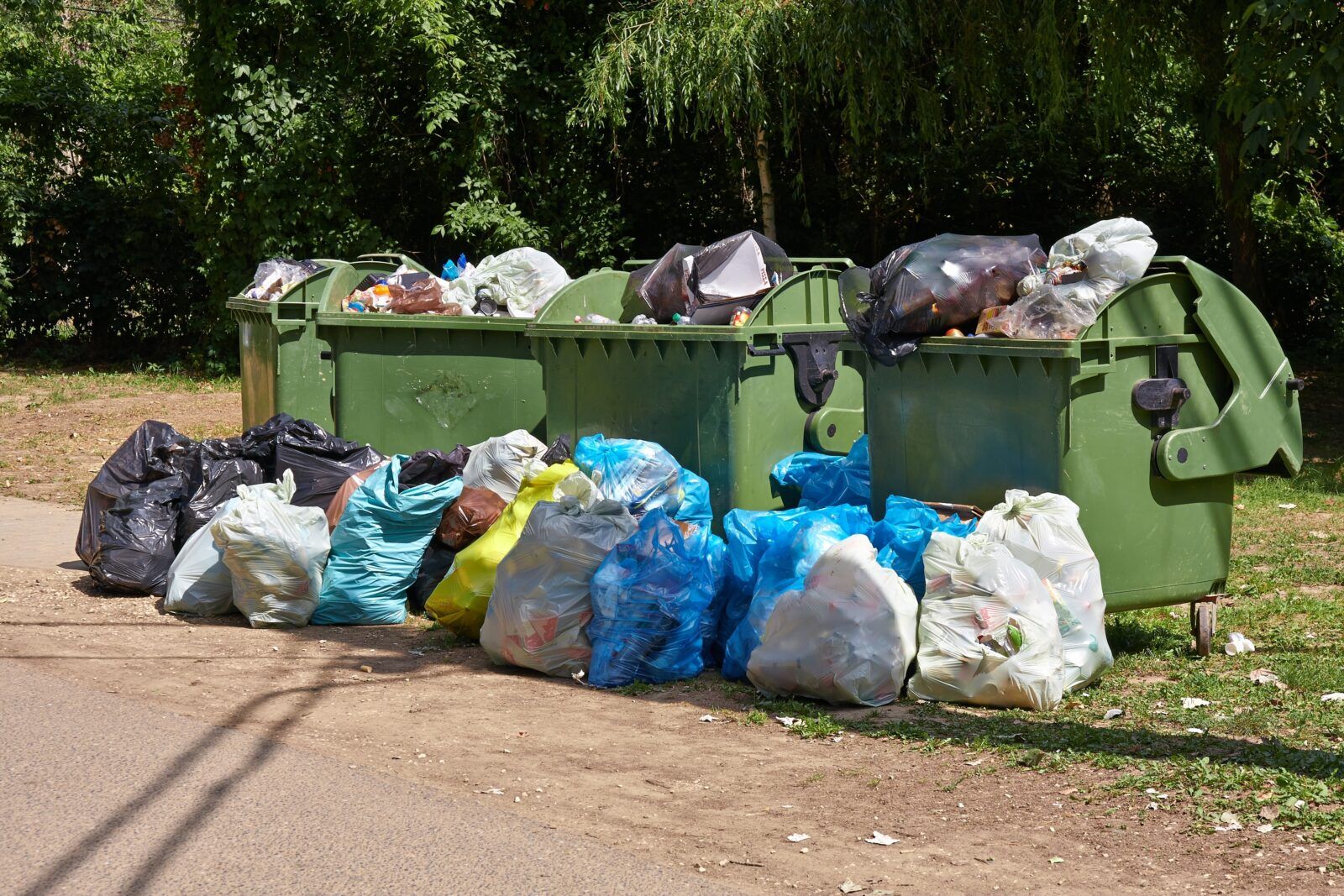 Green trash bins overflowing with garbage, surrounded by colorful bags of trash on the side of a road, in an outdoor setting.